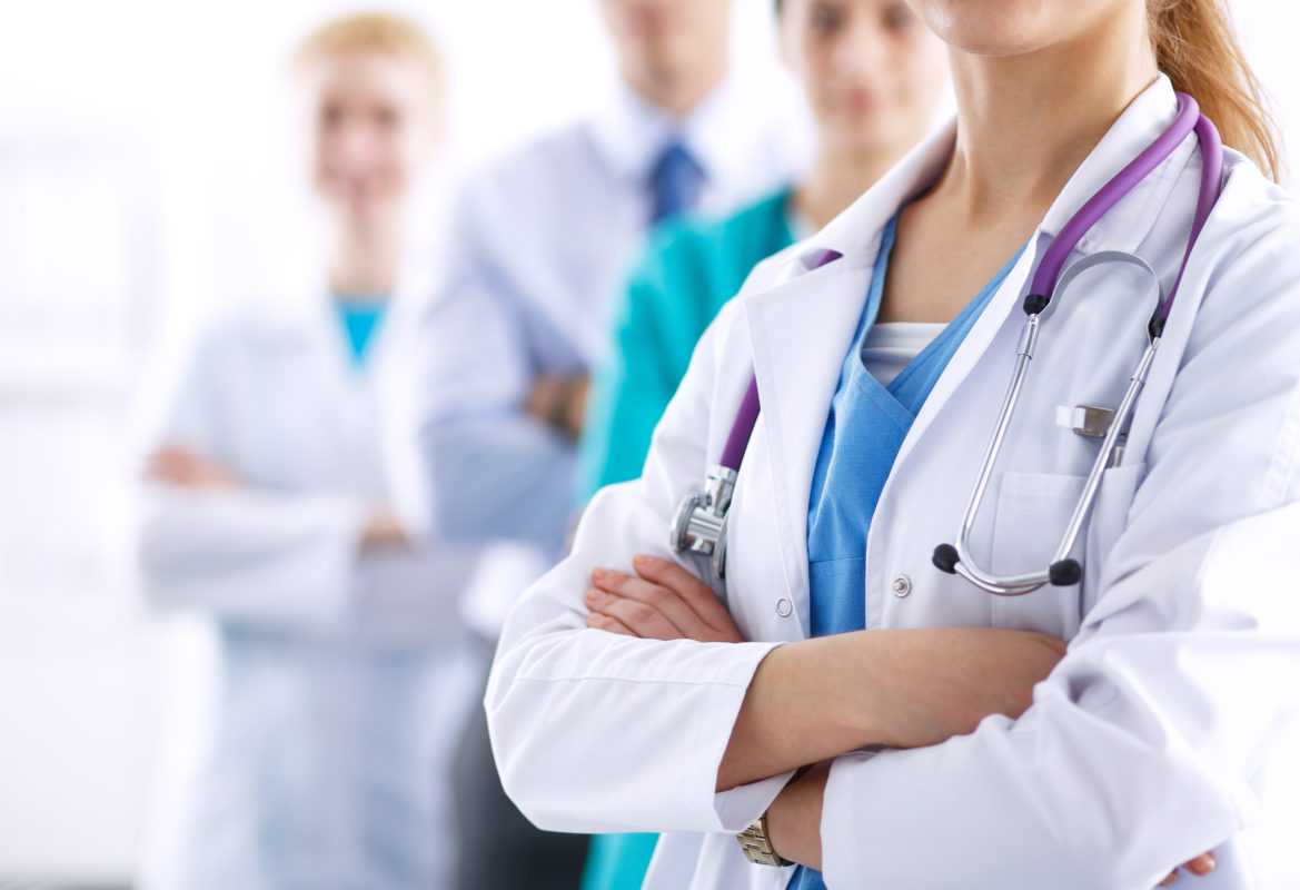 Doctor in white lab coat standing with arms crossed, a purple stethescope draped over her shoulders.